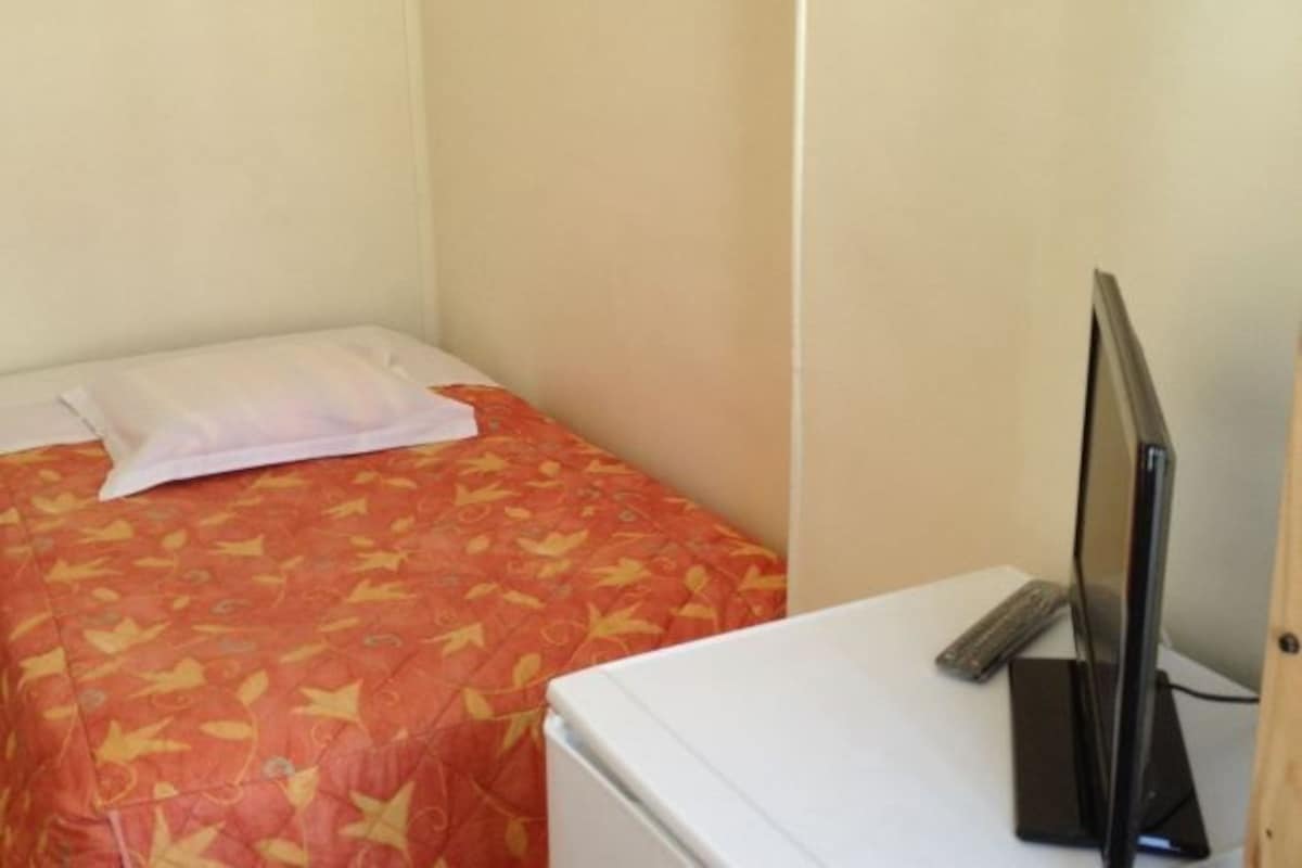 Una habitación pequeña con una cama, un televisor sobre un mueble bajo y un electrodoméstico blanco, probablemente un refrigerador.