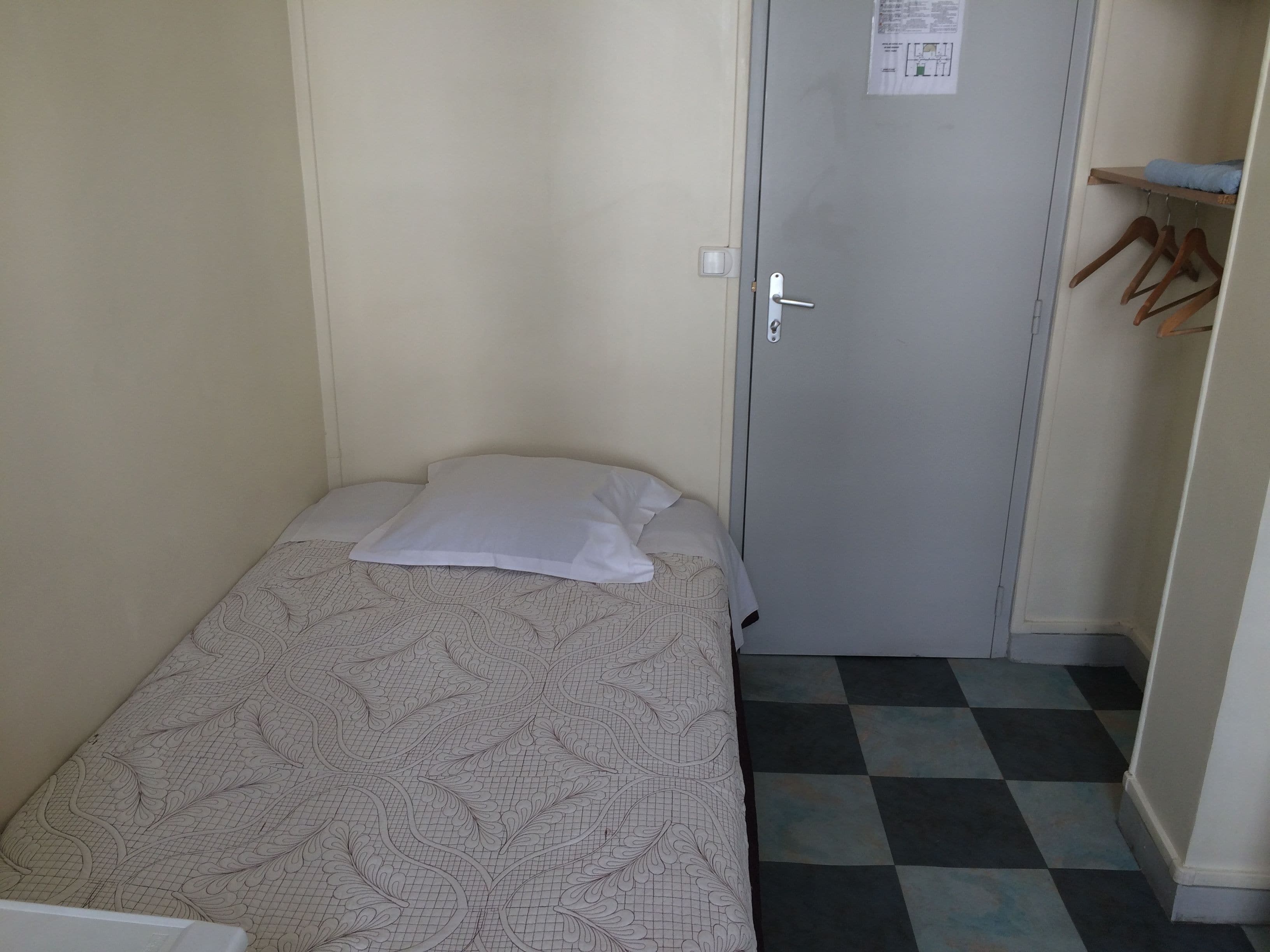 A small, single-bed room with a patterned bedspread, a door with a handle, and a checkered floor.
