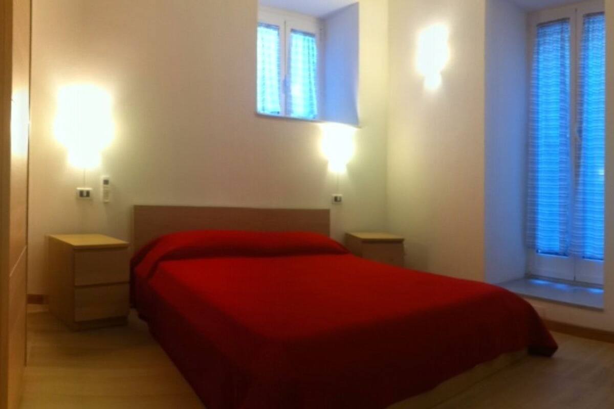 A bedroom with a bed, bedside tables, a desk, and a chair. There are windows with blinds and a wall-mounted TV.