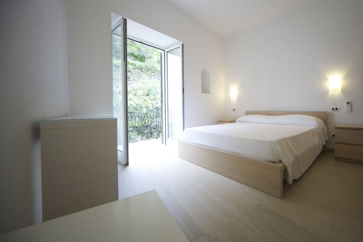 A minimalist bedroom with a large bed, a nightstand, a bedside table, a radiator, and a sliding glass door leading to a balcony.
