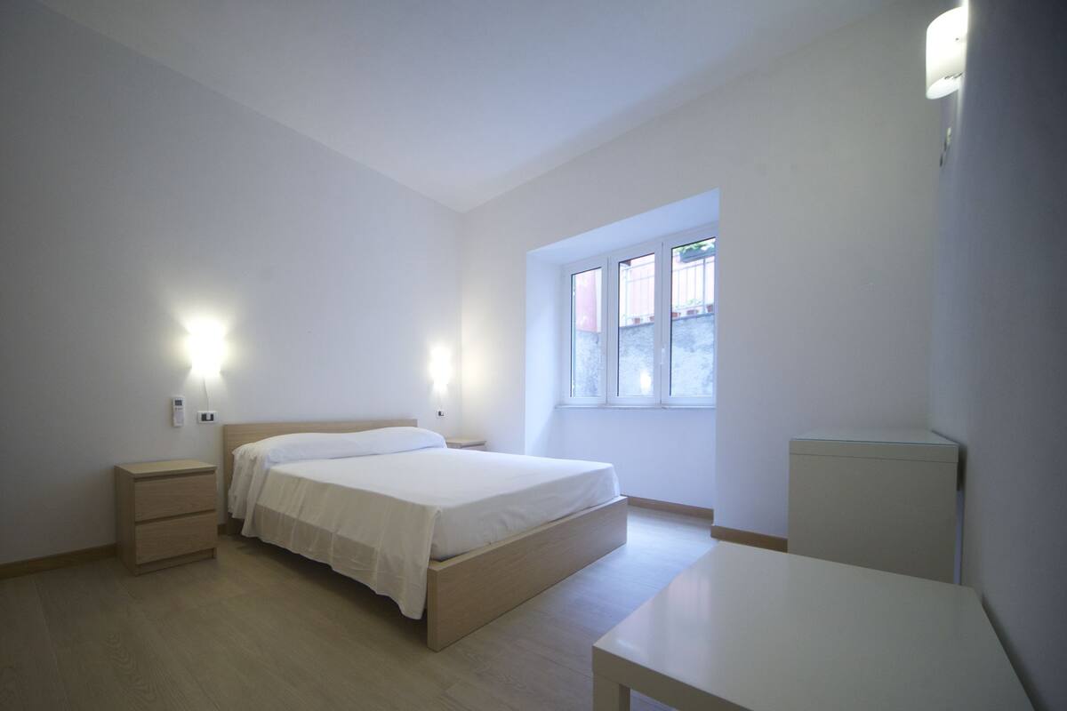 A minimalist bedroom with a single bed, bedside tables, and a window providing natural light.