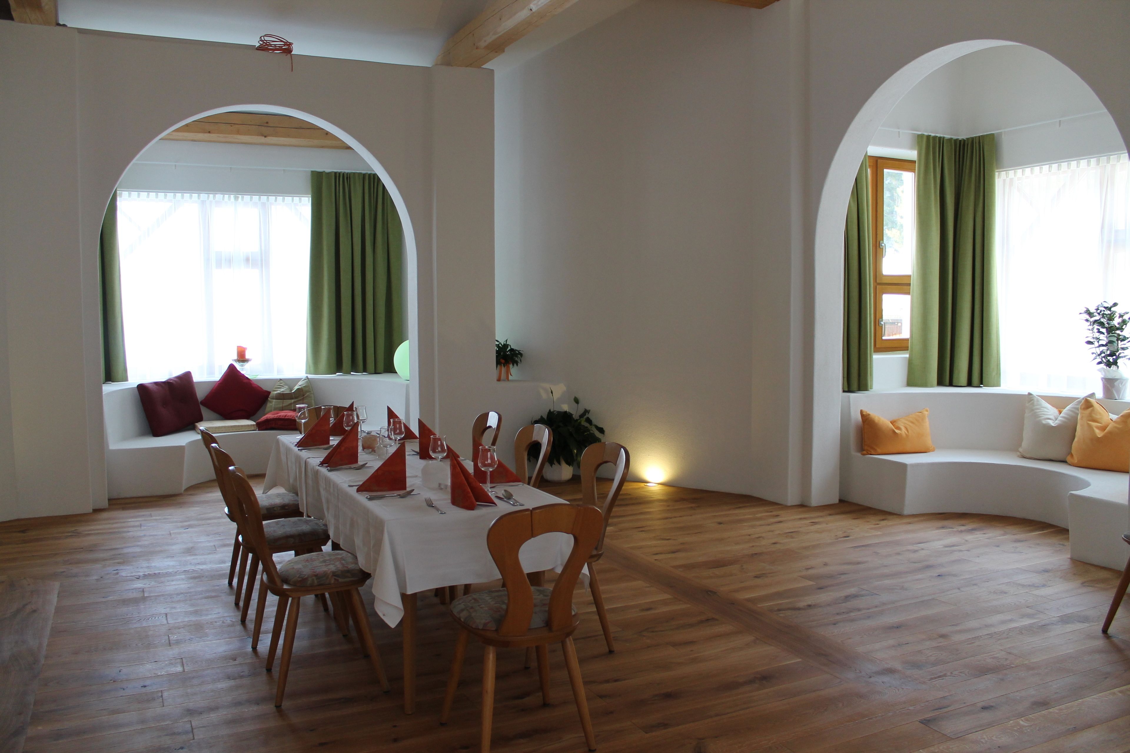 Un comedor con una mesa de madera dispuesta para cuatro personas, rodeada de sillas de madera. En el fondo, un sofá blanco con cojines anaranjados, y una ventana con cortinas verdes.