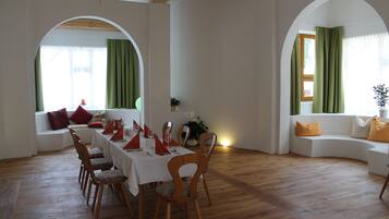 Un comedor con una mesa de madera dispuesta para cuatro personas, rodeada de sillas de madera. En el fondo, un sofá blanco con cojines anaranjados, y una ventana con cortinas verdes.