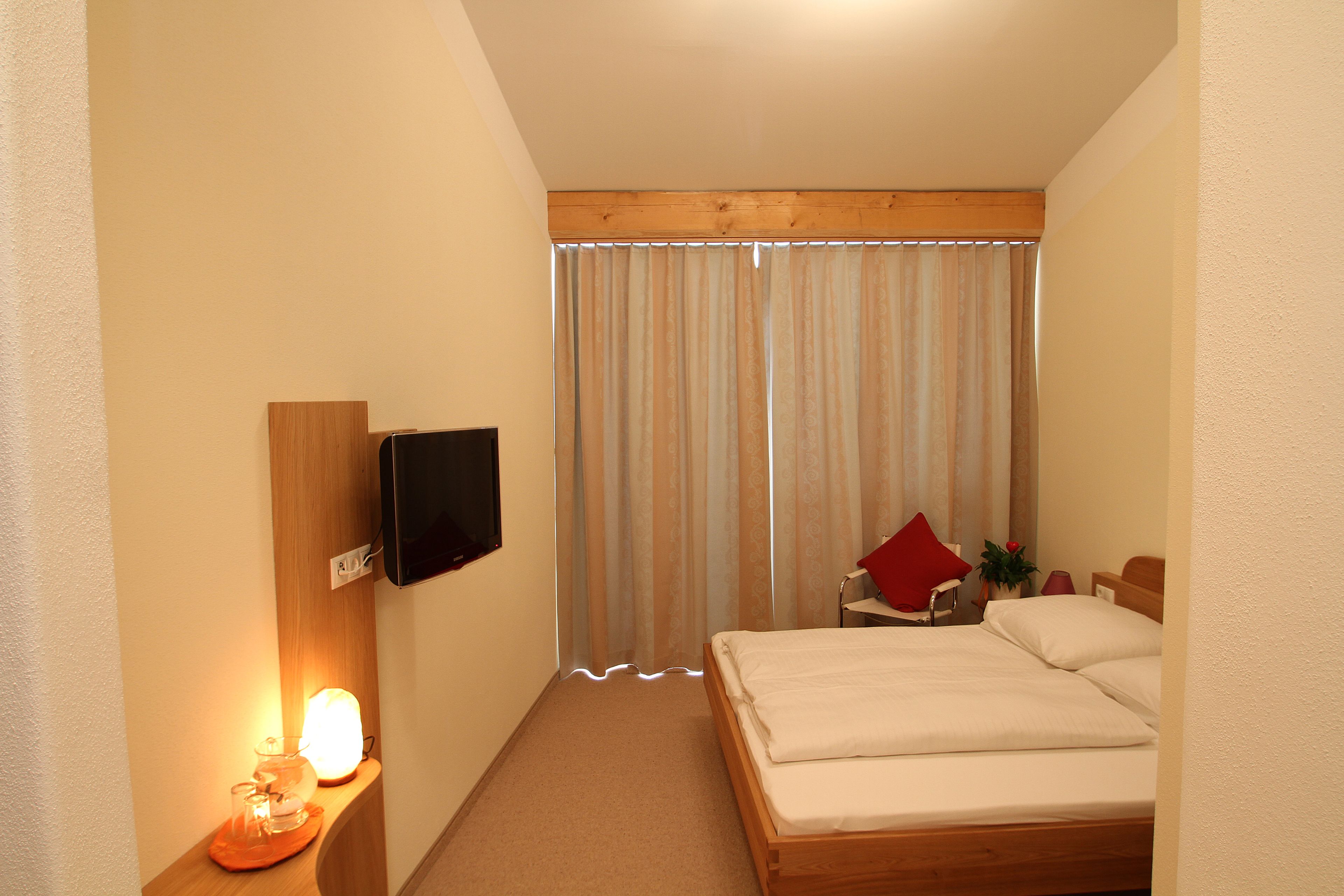 A bedroom with a bed, a TV mounted on the wall, a bedside table with a lamp, and a window with curtains.