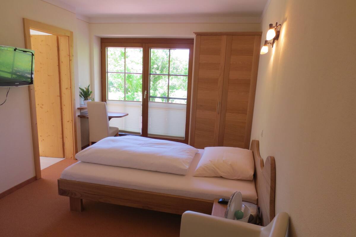 A bedroom with a bed, a chair, a small table, a TV, and a window with a view of trees.