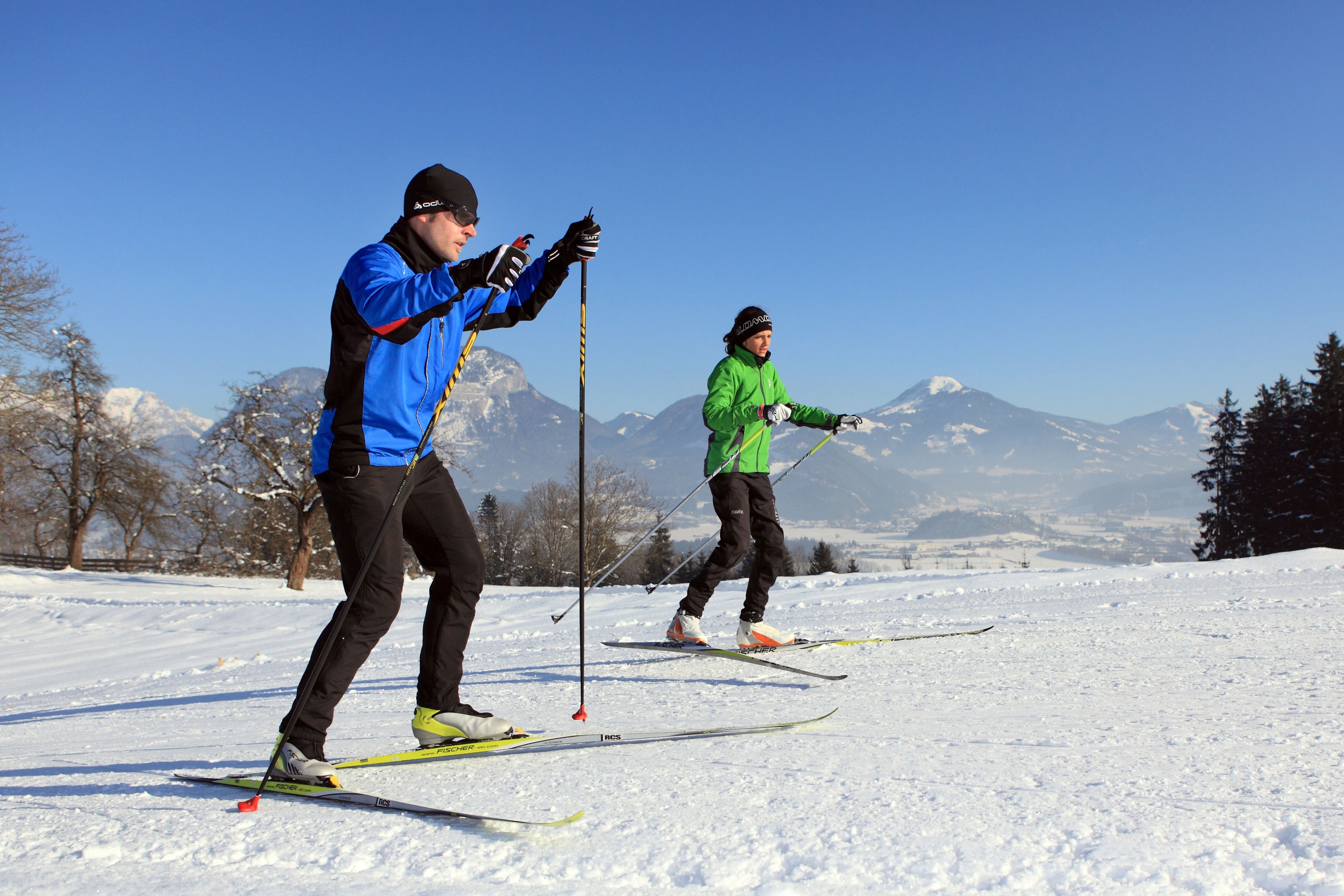 Two people cross-country skiing on a snowy landscape with mountains in the background.