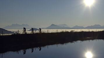 A serene lakeside scene with cyclists and a bench, surrounded by mountains and a clear sky.