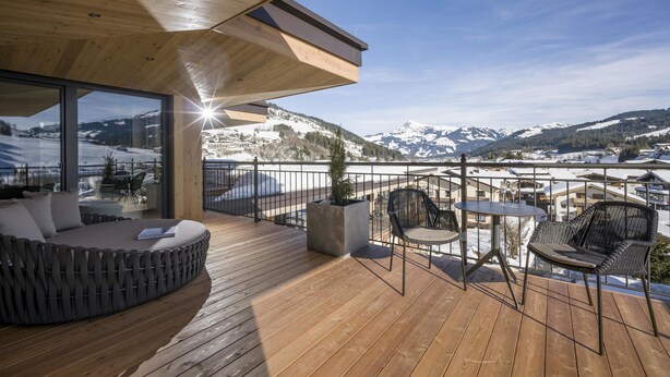 Una terraza de madera con zona de estar, una mesa y sillas, con vistas a un paisaje montañoso nevado.