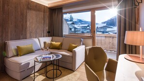 Una habitación de hotel moderna con una pared de acento de madera, un sofá, una silla, una mesita con una revista y un ventanal con vistas a casas y montañas nevadas.