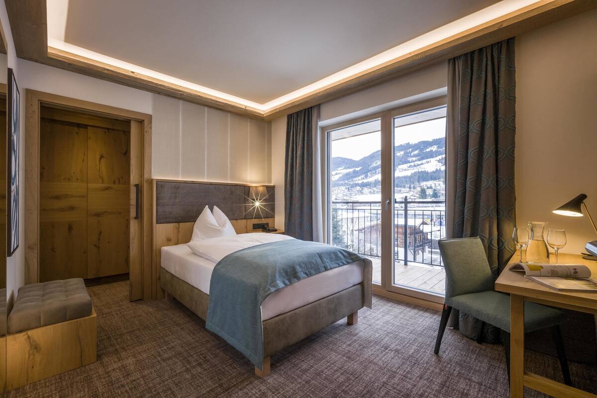 Ein Hotelzimmer mit Bett, Schreibtisch, Stuhl, Fenster mit Blick auf eine verschneite Landschaft und einem Balkon.