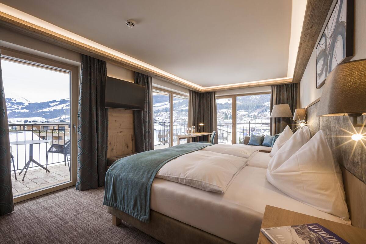 Ein Hotelzimmer mit einem großen Bett, einem Schreibtisch, einem Stuhl und einem Balkon mit Blick auf schneebedeckte Berge.