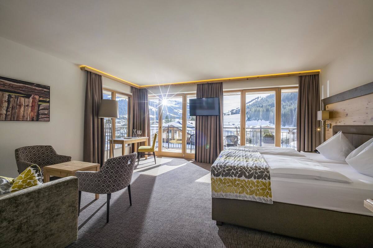 Ein Hotelzimmer mit einem großen Bett, einem Schreibtisch, einem Sessel und einem Fernseher. Ein Fenster bietet Blick auf die Berge.