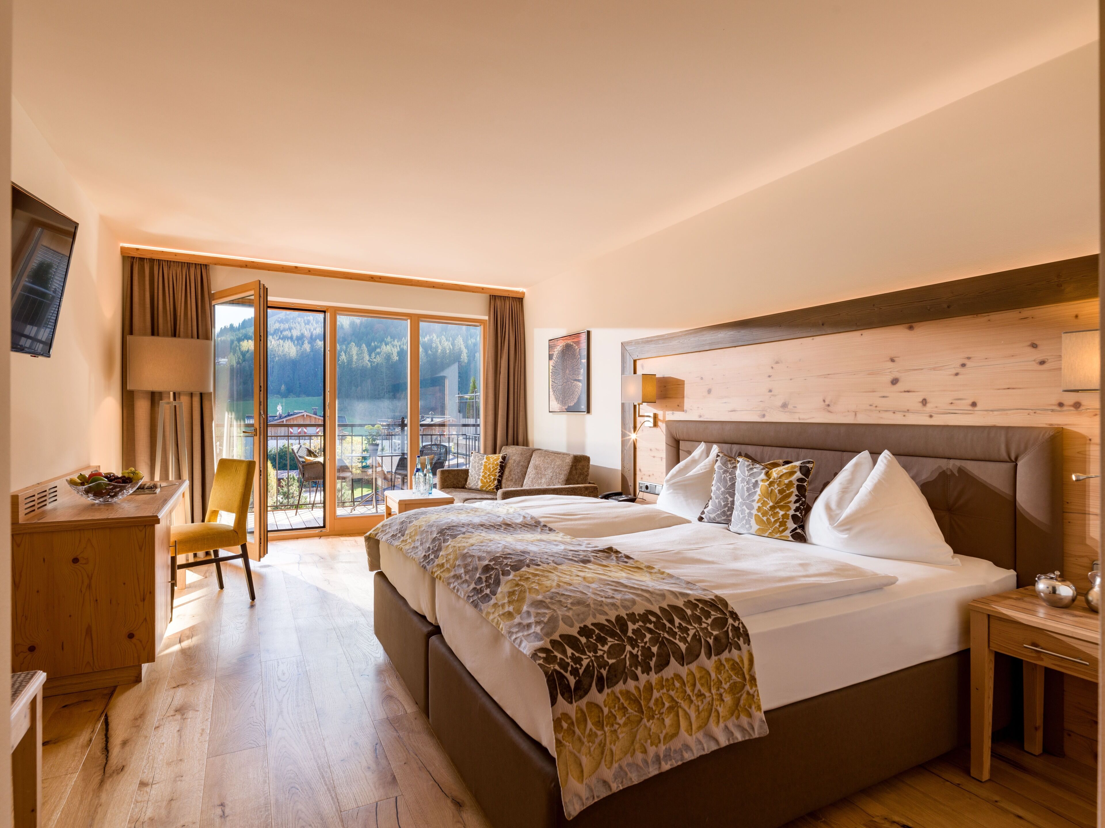 A hotel room with a large bed, a desk, a chair, and a balcony with a view of the mountains.