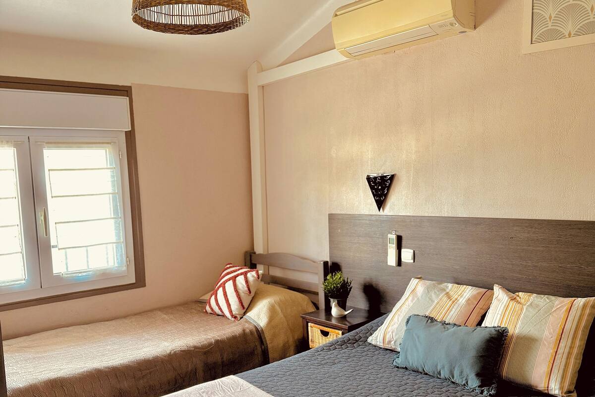 A bedroom with two beds, a hanging light fixture, a bedside table with a plant, and a wall-mounted air conditioner.
