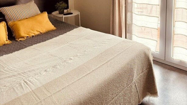 Un dormitorio con cama, mesita de noche, luz de pared y vistas a través de una ventana con cortinas.