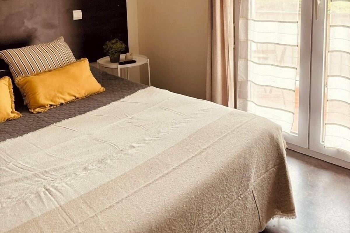 A bedroom with a bed, a nightstand, a wall-mounted light, and a view through a window with curtains.