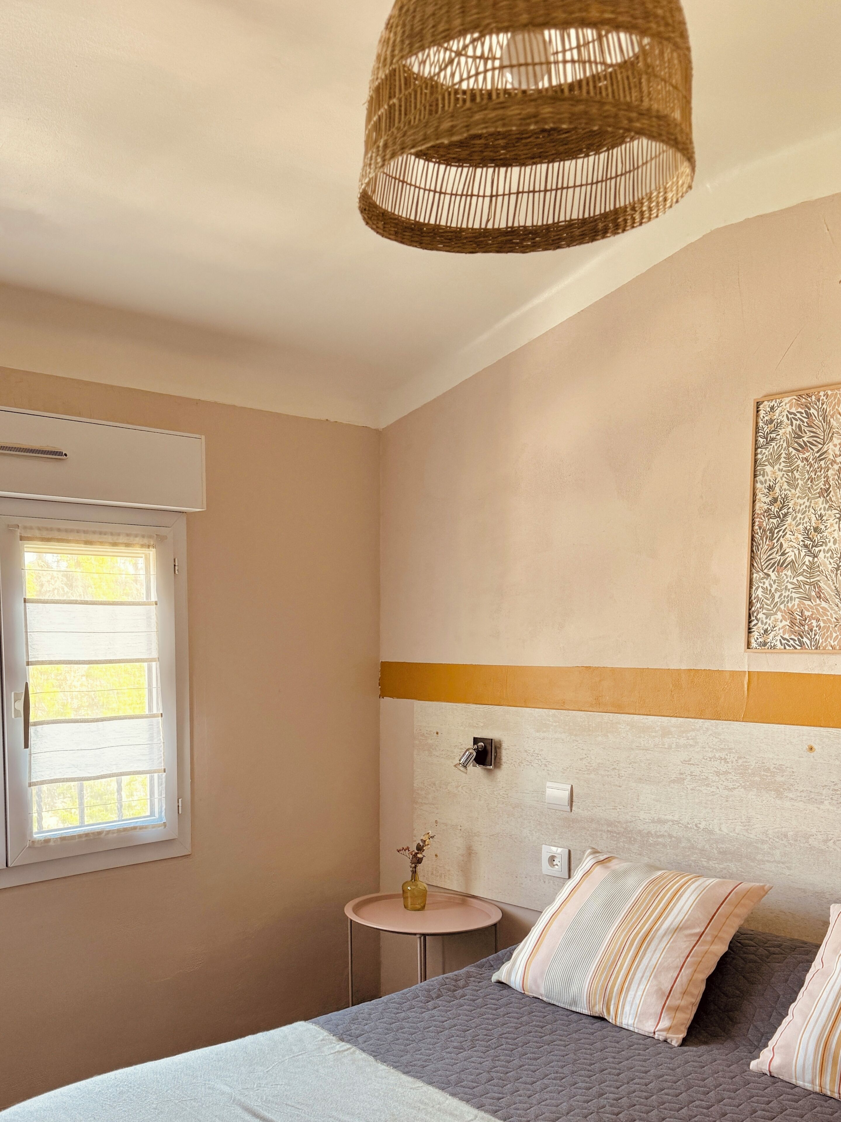Ein Schlafzimmer mit Bett, Nachttisch, Pendelleuchte, Fenster und einem an der Wand befestigten Kunstwerk.