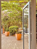 On aperçoit, à travers une porte vitrée, une cour intérieure agrémentée de deux grands pots de plantes, d’une zone pavée et d’un portail en métal.