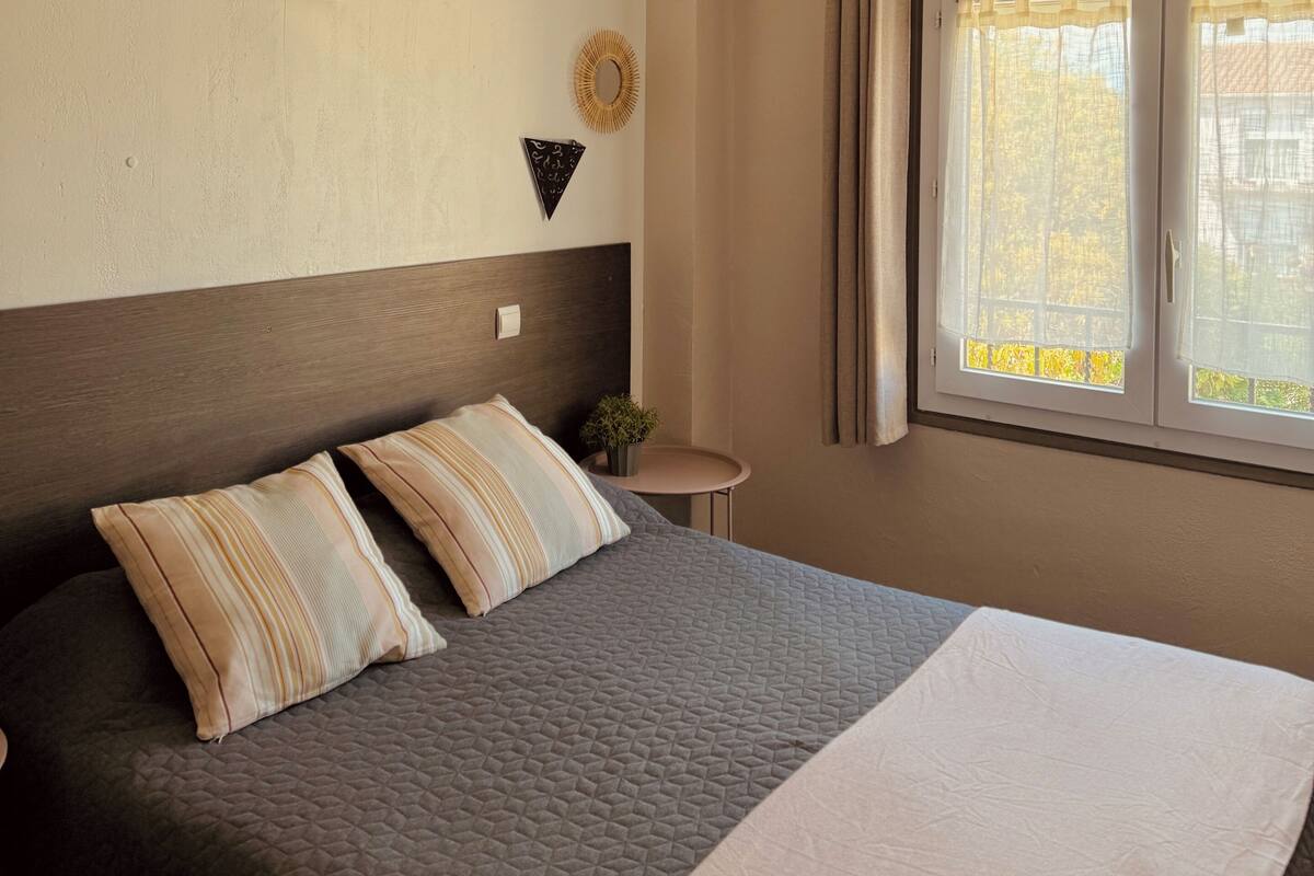 A bedroom with a large bed, a hanging wicker light fixture, a round mirror, and a window with curtains.