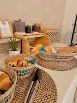 Une table de petit-déjeuner avec des paniers en osier contenant des viennoiseries et du pain, un menu encadré et une petite plante en pot.