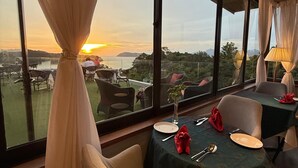 A dining area with a view of the sunset over a body of water, large windows, and a table set for two. - Ambong Rainforest Retreat (Langkawi)