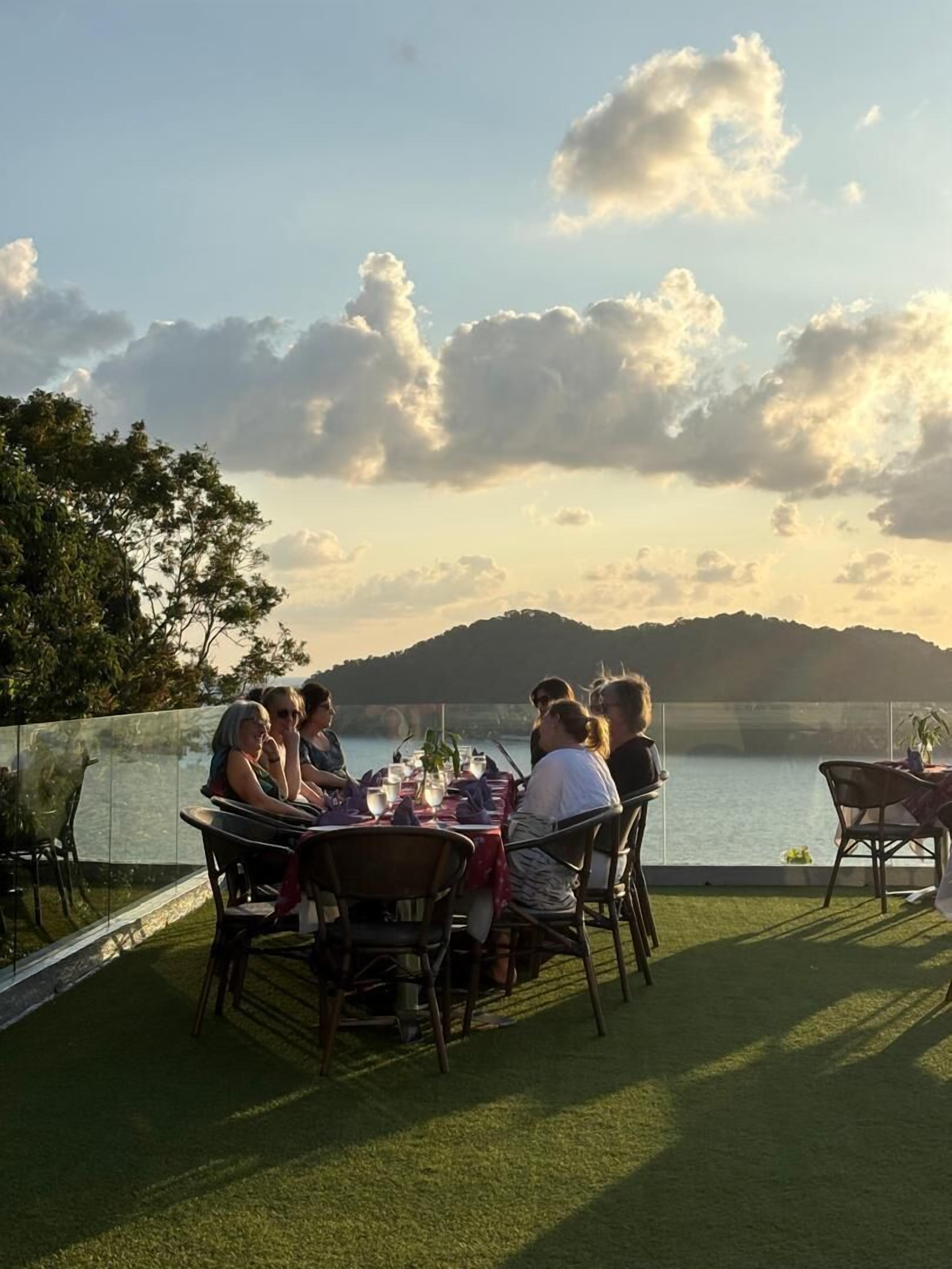 Een groep mensen dineert aan een buitentafel met uitzicht op een meer en bergen.
