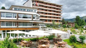 Ein Hotel mit mehreren Stockwerken, einer Außenbereichs-Sitzgelegenheit mit Sonnenschirmen und einer Bergkulisse.