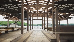 A wooden deck with benches, a covered area with a roof, and a view of a body of water and greenery in the background. - Hiogiso (Shima)