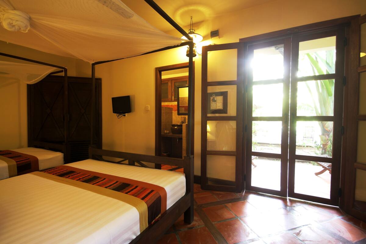 Habitación de hotel con dos camas, estructura de dosel sobre la cama, televisor y puerta corrediza de vidrio que da a un espacio exterior.