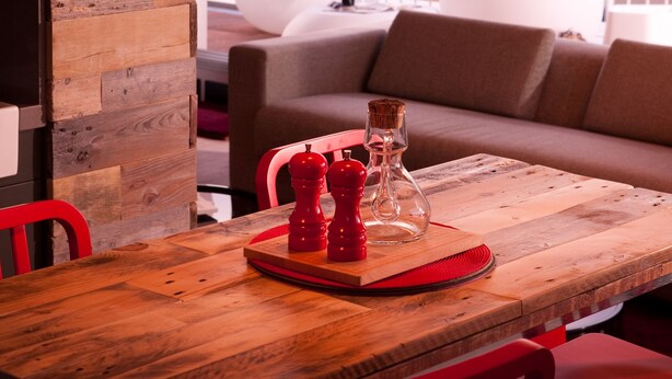 Een houten eettafel met rode stoelen, een rode pepermolen en een glazen karaf. Op de achtergrond is een witte bank en een gestreept kussen zichtbaar.