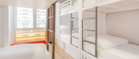 A dormitory room with bunk beds, a window with a view of buildings, and a wooden floor.