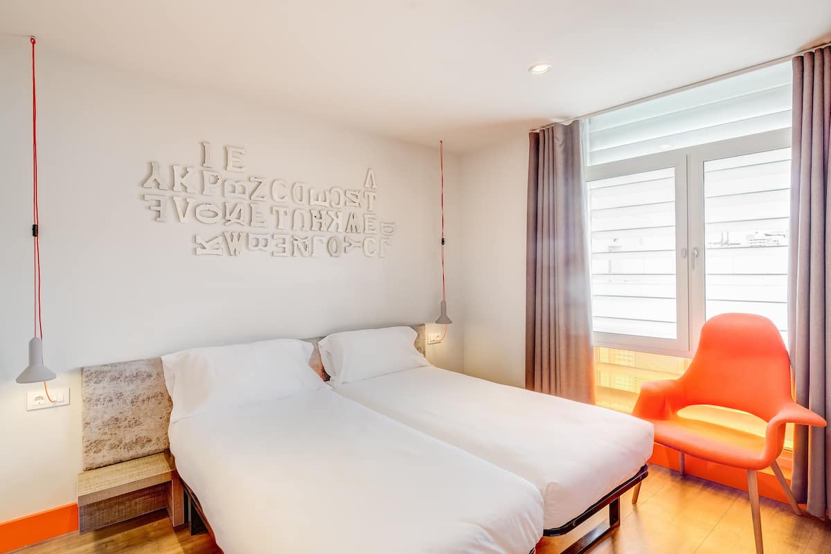 A modern bedroom with a large bed, a bright window, and a red chair.