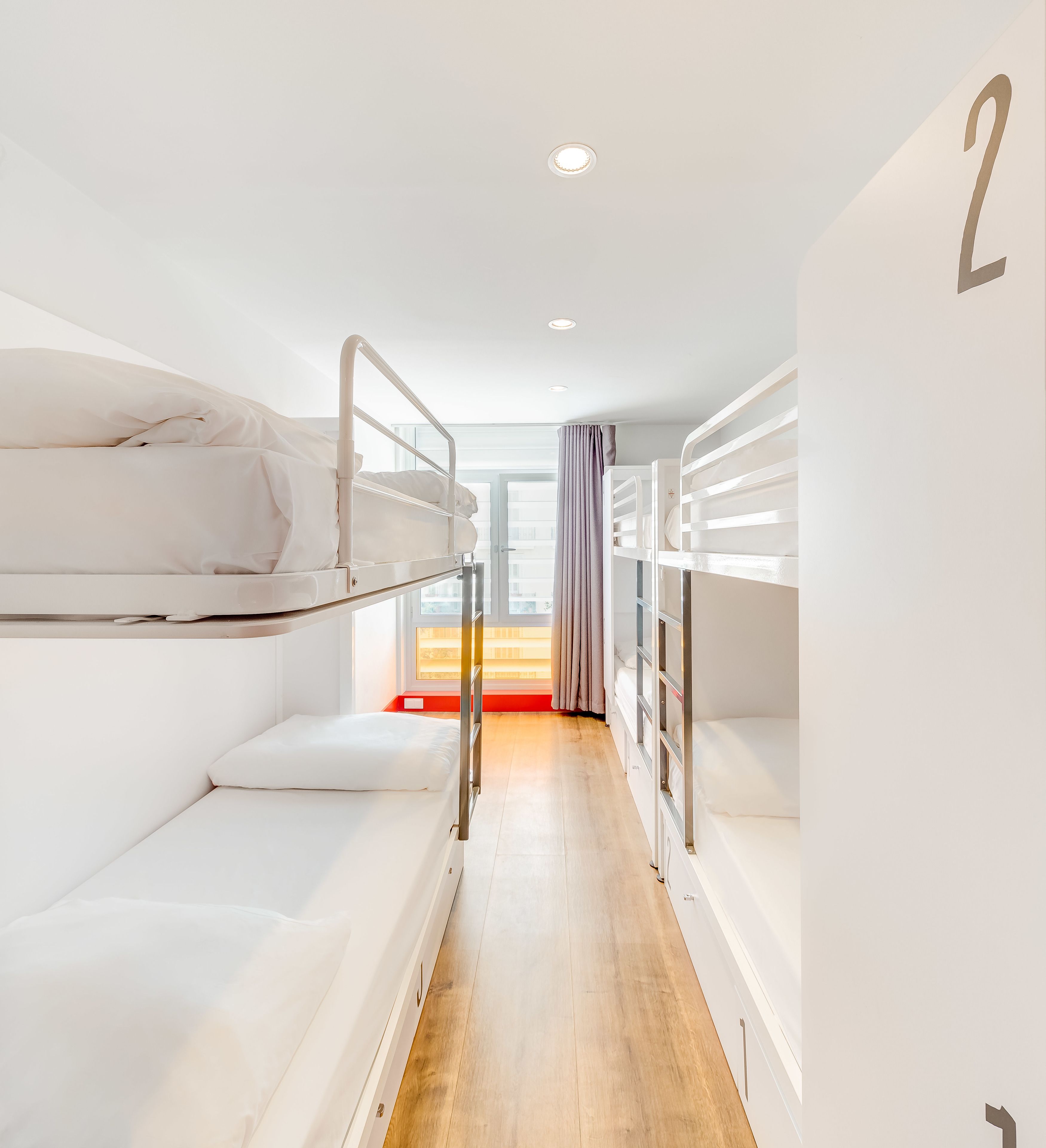 Un estrecho pasillo con literas a ambos lados, suelo de madera y una ventana con cortinas.