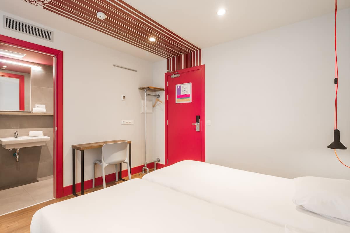 A hotel room with a bed, a desk, a chair, a sink, and a red door.