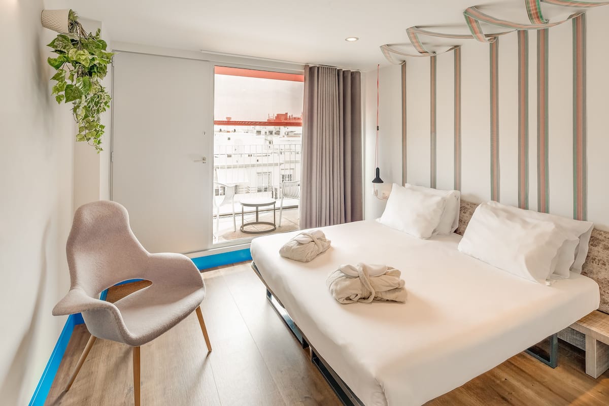 A modern hotel room with a large bed, a chair, and a view of the city through a glass door.