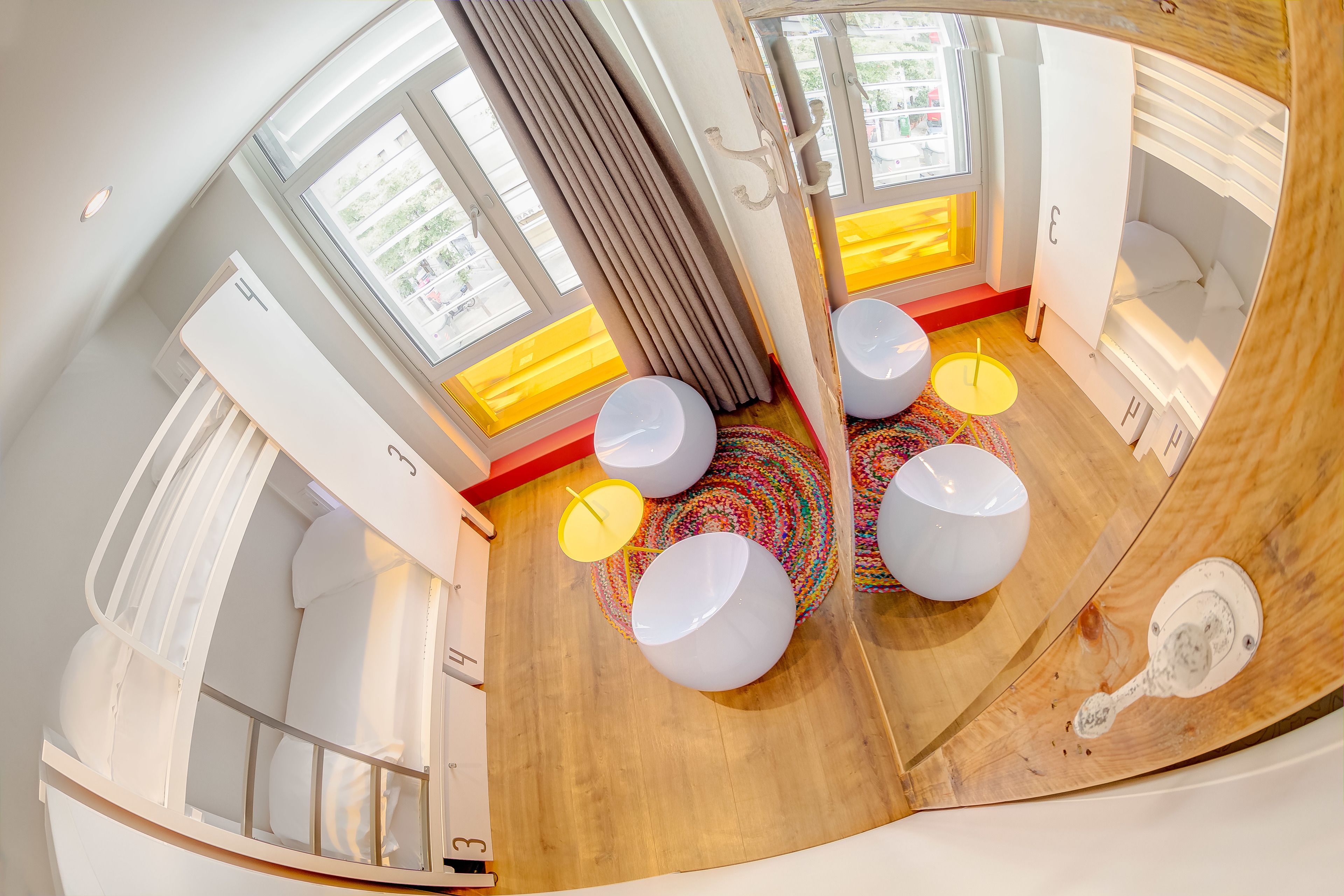 Una habitación moderna con suelo de madera, paredes blancas y un ventanal con cortinas. Seis mesas blancas y redondas con tapas amarillas y una alfombra tejida de colores.