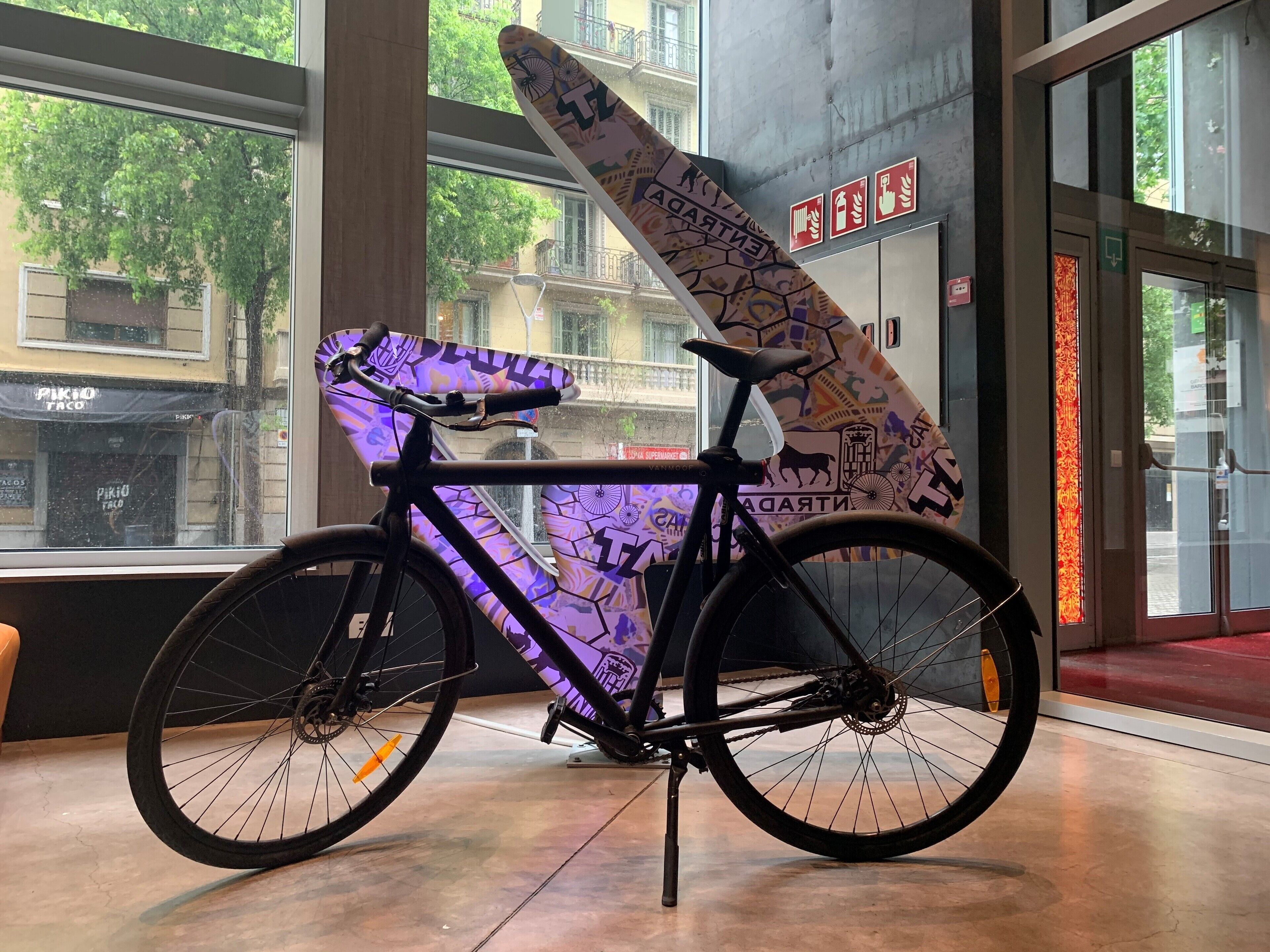 A bicycle with a colorful, patterned seat and a large, decorated front wheel, leaning against a wall with large windows and a view of buildings outside.