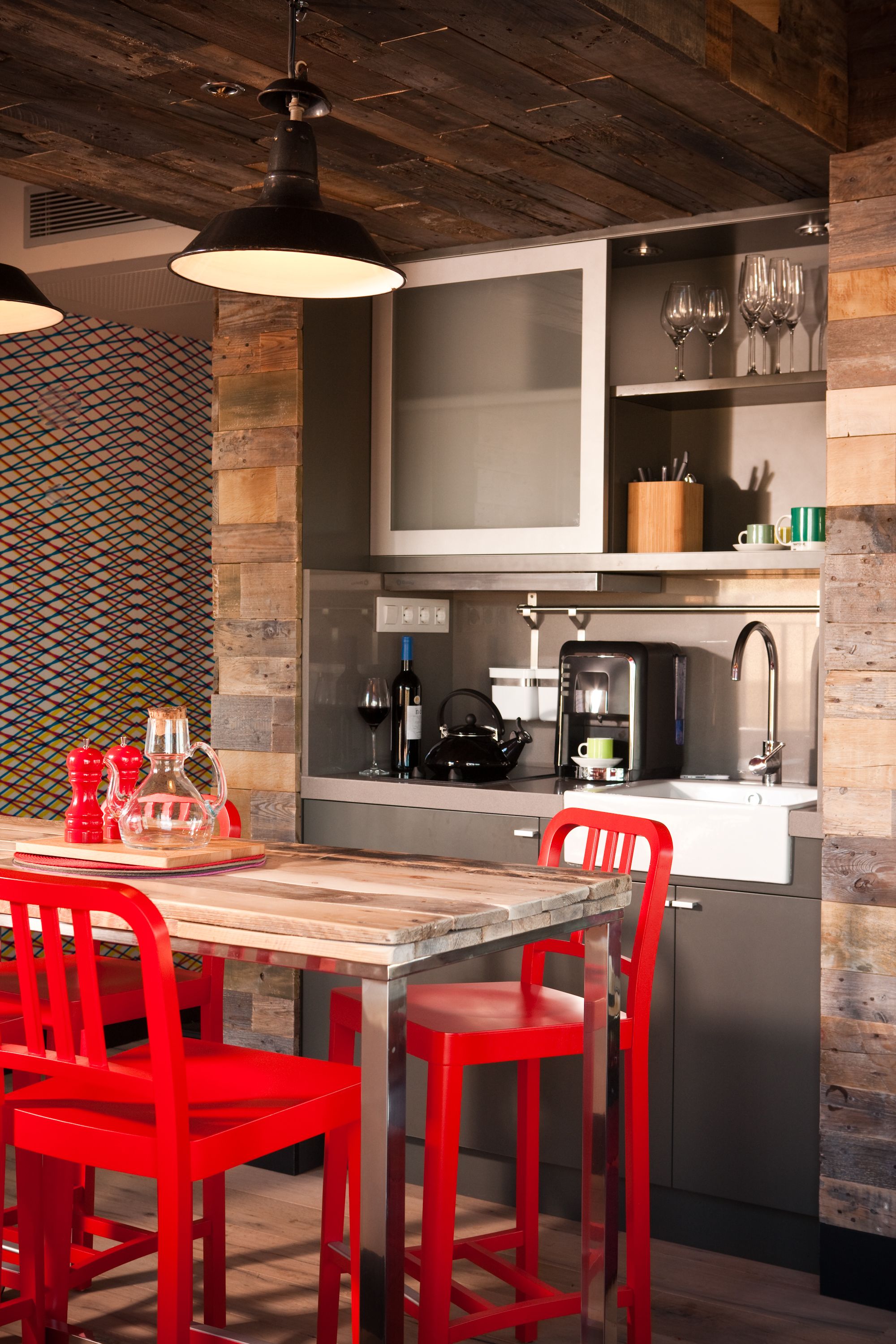 A modern kitchen with a wooden table and red chairs, a coffee machine, and wine glasses on the counter.