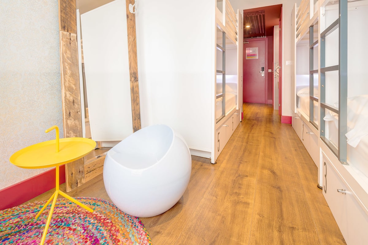A modern bathroom with a wooden floor, a white bathtub, a yellow stool, and a colorful rug.