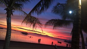 A beach scene with palm trees, boats, and people silhouetted against a sunset. - Palms Cove Resort (Panglao)