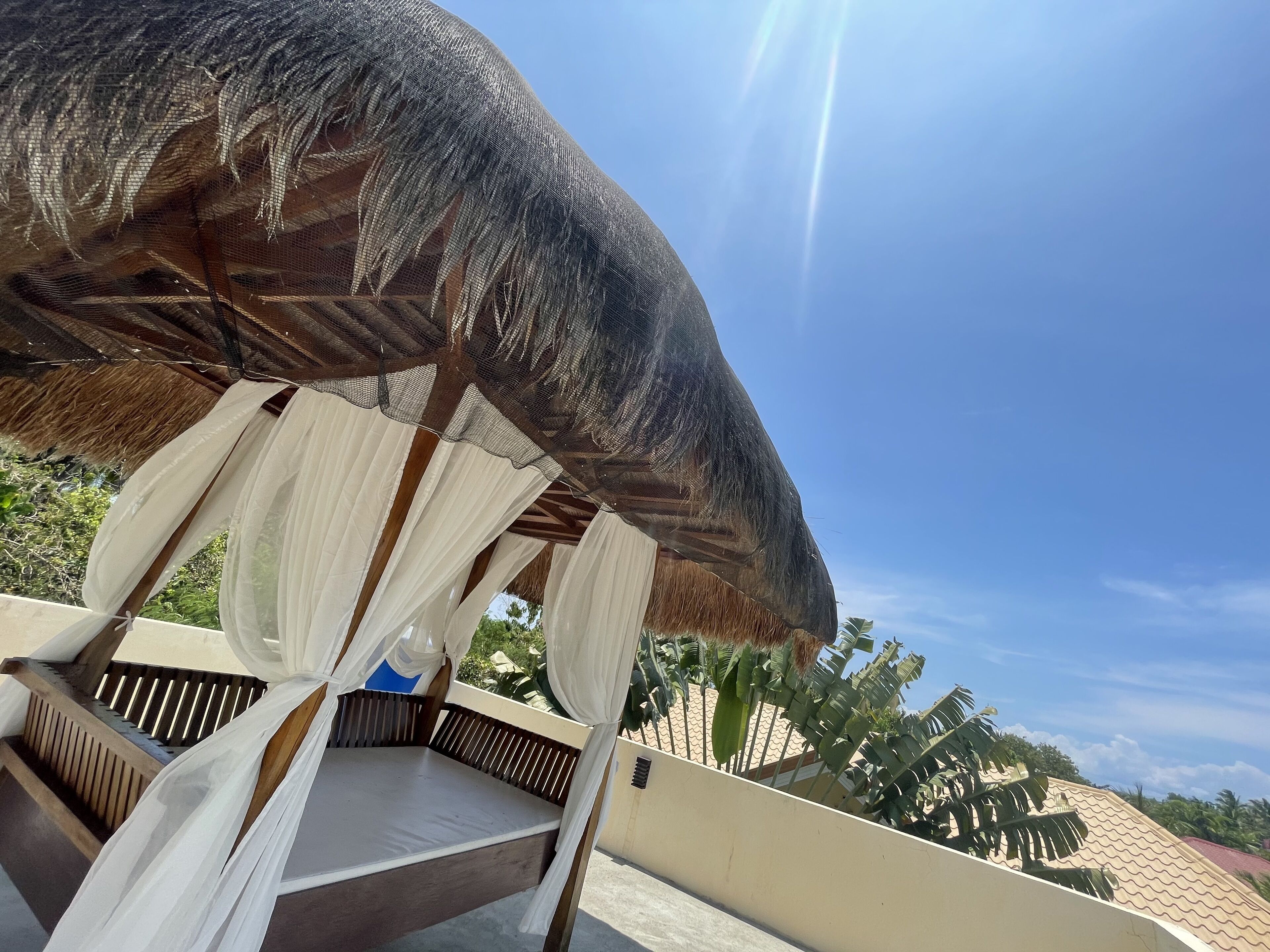 A bed under a thatched roof with white curtains, a view of palm trees, and a clear blue sky.