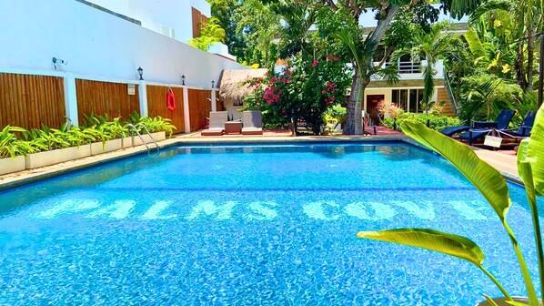 Una piscina con las palabras 'PALMS CONVOY' escritas sobre el agua, rodeada de follaje y sillas de descanso.