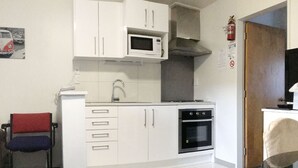 A compact kitchen with white cabinets, a microwave, an oven, and a sink. There is a chair with a red cushion and a towel hanging on the door. - Adelaide Motel (Wellington)