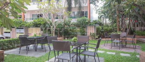 An outdoor patio area with wicker furniture and a small table, surrounded by greenery and a building in the background.