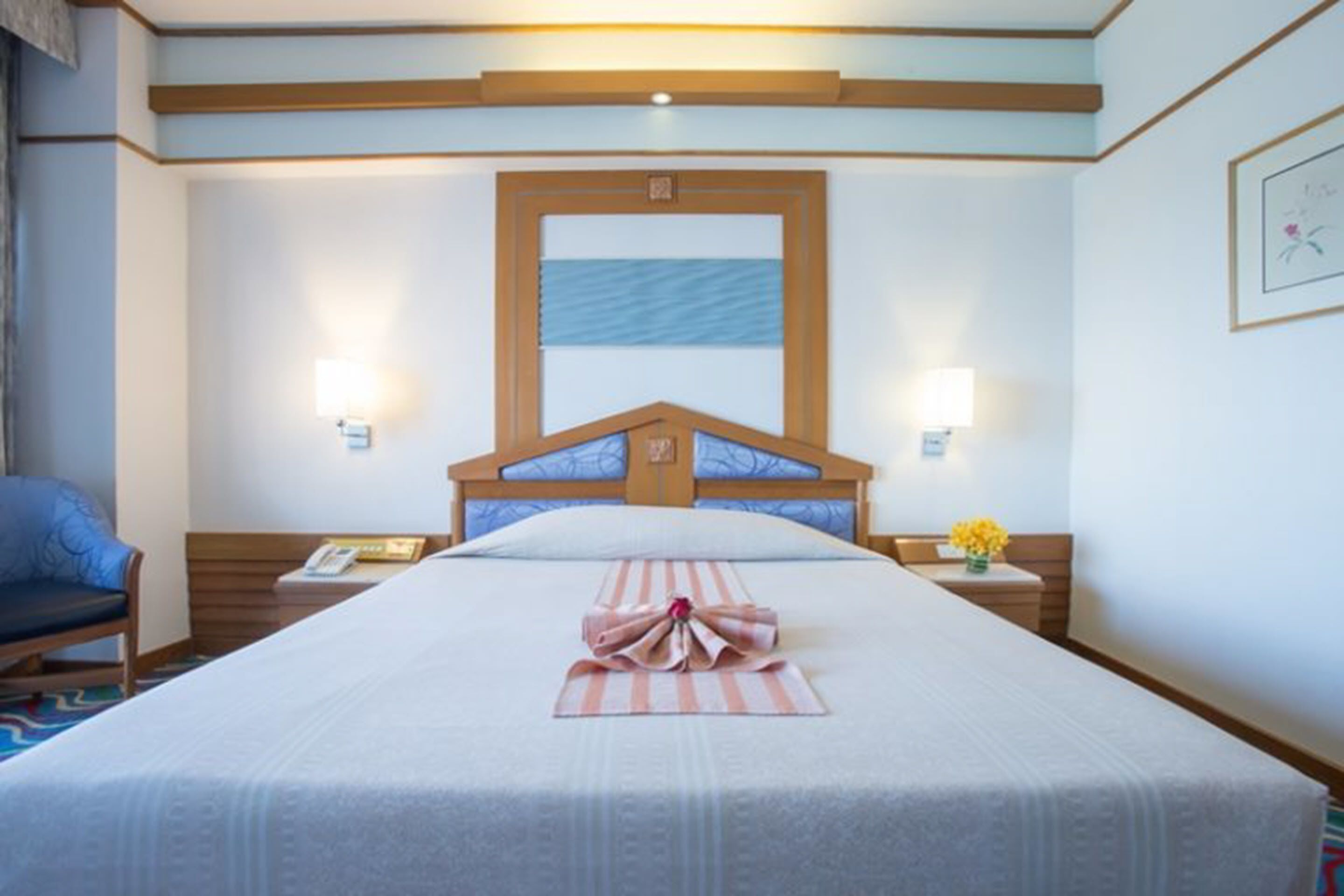 A hotel room with a large bed, wooden headboard, two bedside tables with lamps, a chair, and a framed picture on the wall.