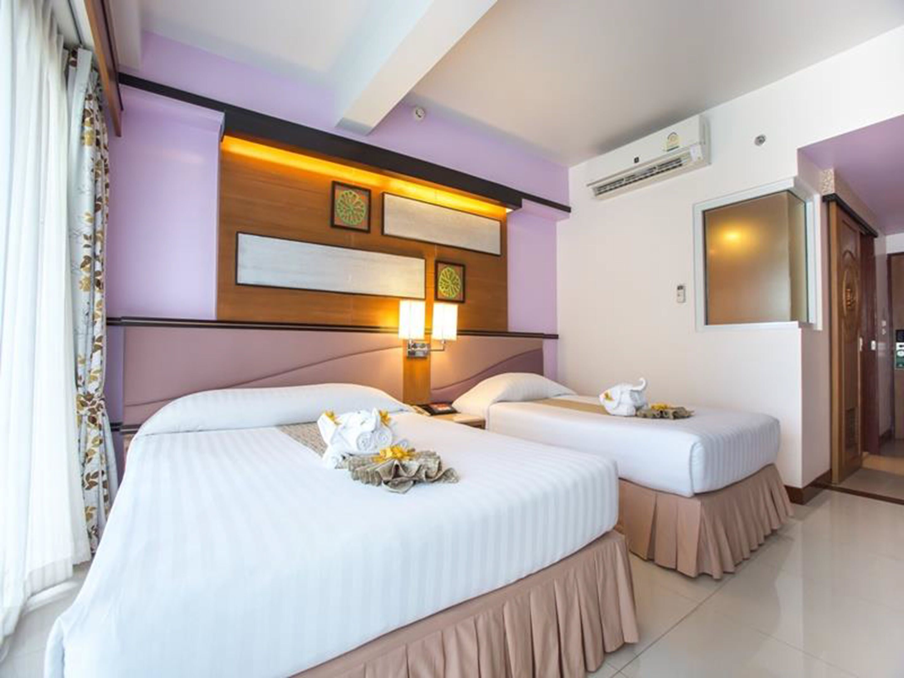 A hotel room with two beds, a headboard with a light fixture, a wall-mounted air conditioner, and a door leading to another room.
