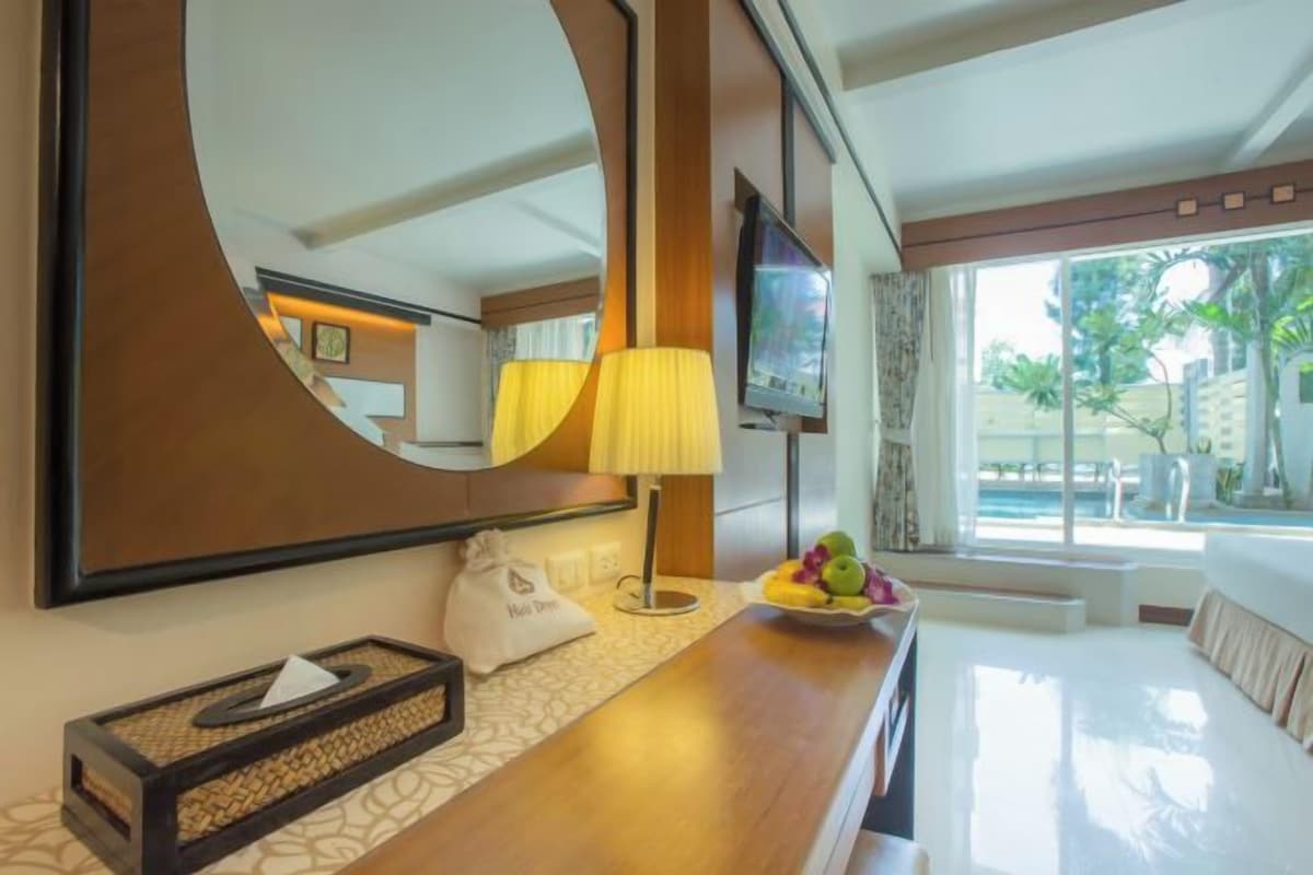 A hotel room with a large mirror, a wooden side table with a bowl of fruit, a lamp, and a television.