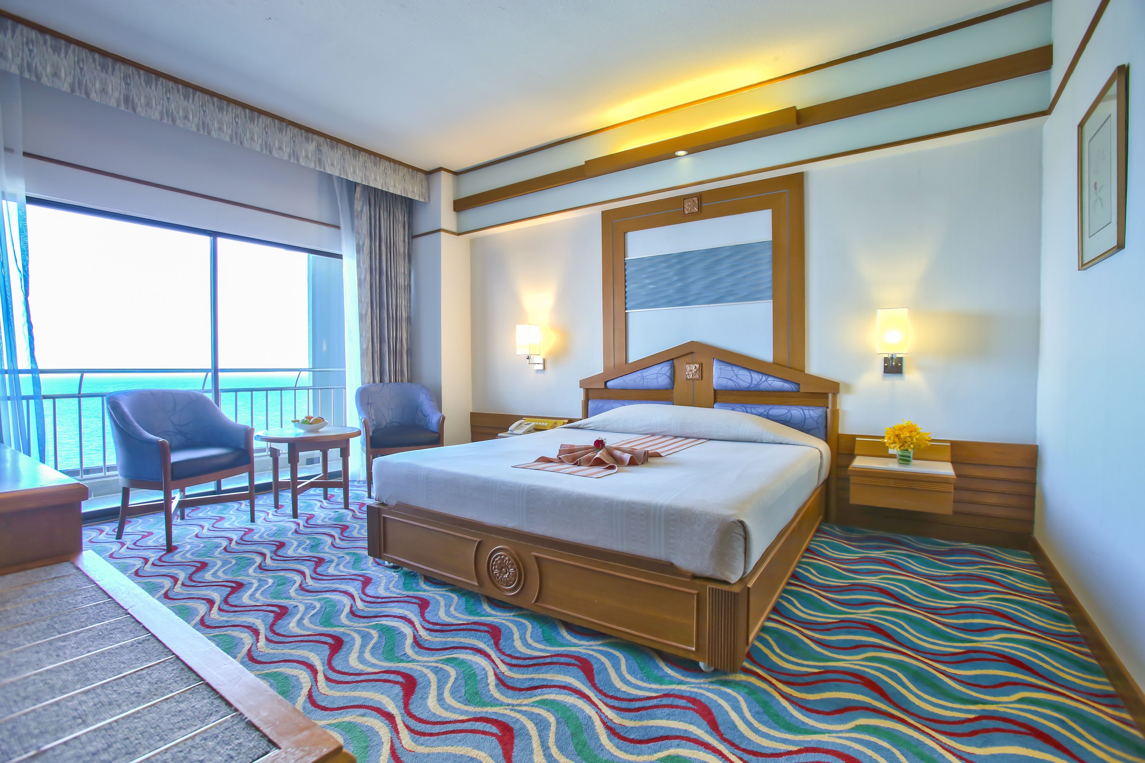 Une chambre d’hôtel avec un grand lit, une chaise, une petite table, une table de chevet avec une lampe, et une vue sur la mer par de grandes fenêtres.