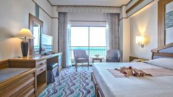 Habitación de hotel con cama, escritorio con lámpara, silla, televisor y vista al océano a través de amplias ventanas.
