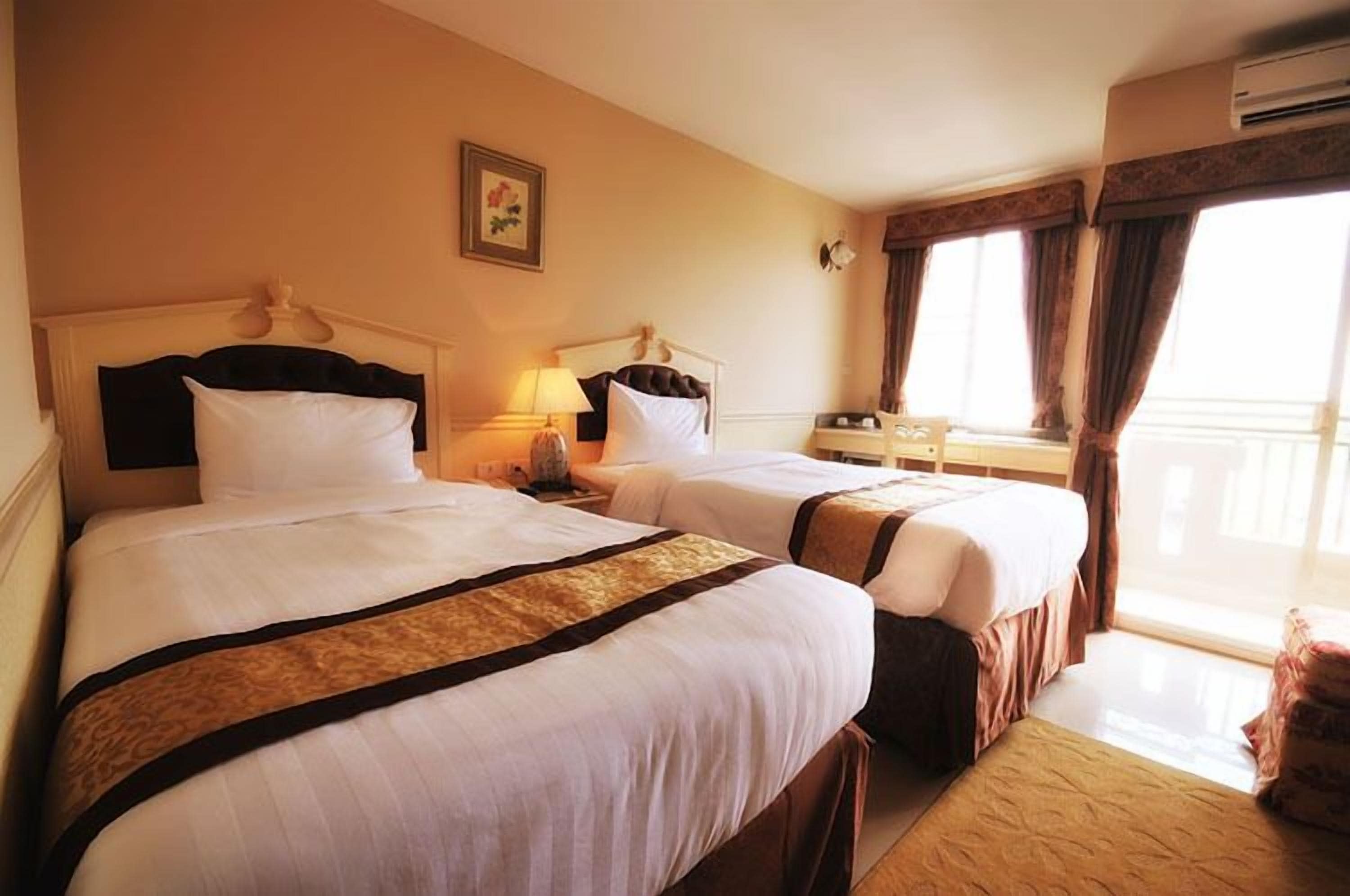 Quarto de hotel com duas camas, uma mesinha de cabeceira com abajur, uma janela com cortinas e ar-condicionado fixado na parede.